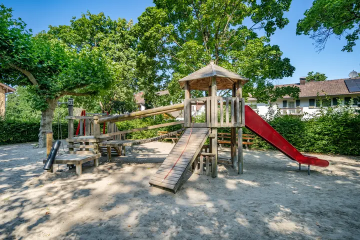Holzspielturm mit roter Rutsche auf einem sonnigen Sandspielplatz unter Bäumen